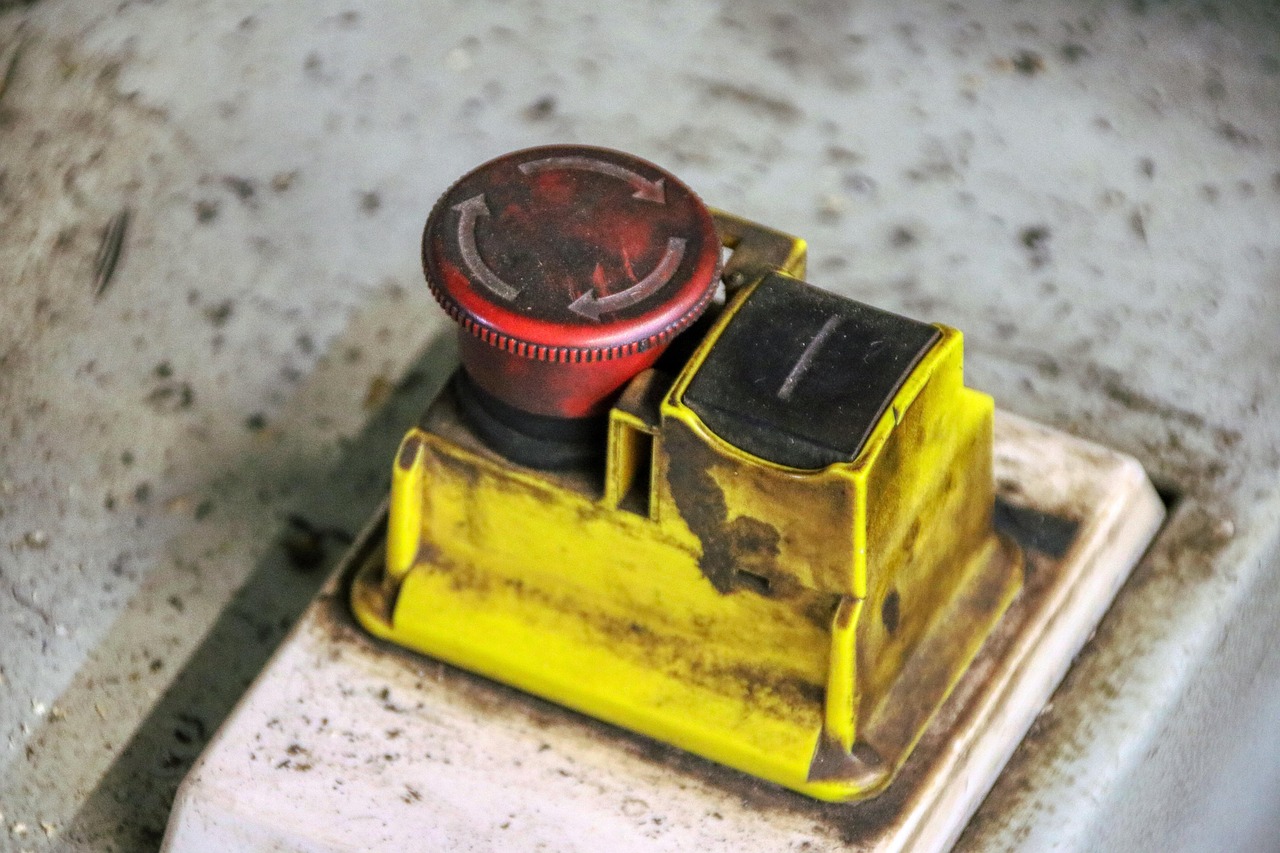 A worn red mushroom-head emergency stop button on a yellow housing, beside a black rocker switch, mounted on a machine