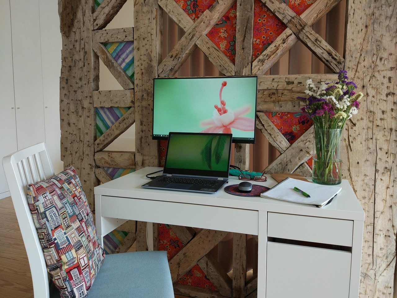 A tidy white home-office desk with a laptop connected to an external monitor, a chair and a vase of dried flowers