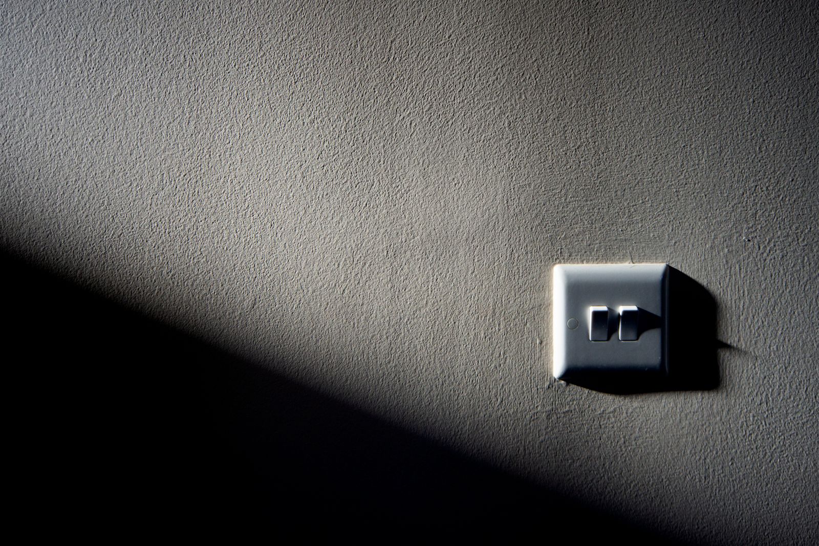 A rocker light switch on a rough painted wall, lit from the upper right so that half the wall is bright and a hard diagonal shadow covers the lower left corner