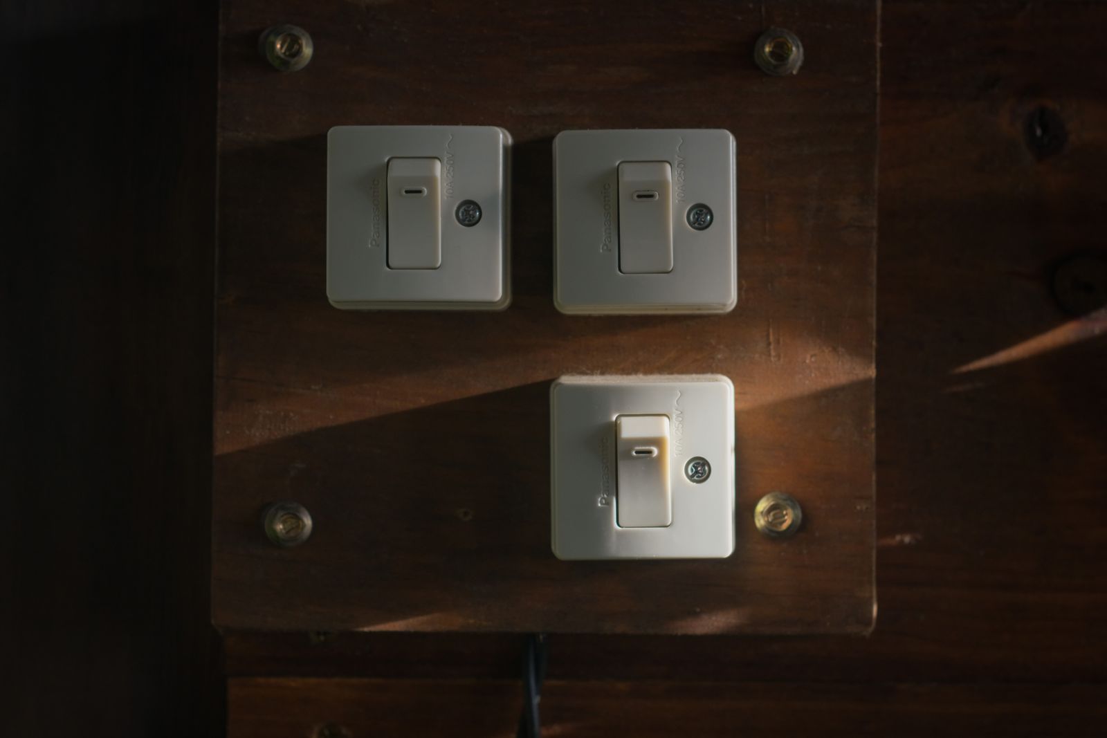 Three white rocker light switches mounted on a dark polished wooden panel, two side by side above and one below, a shaft of daylight falling across the wood