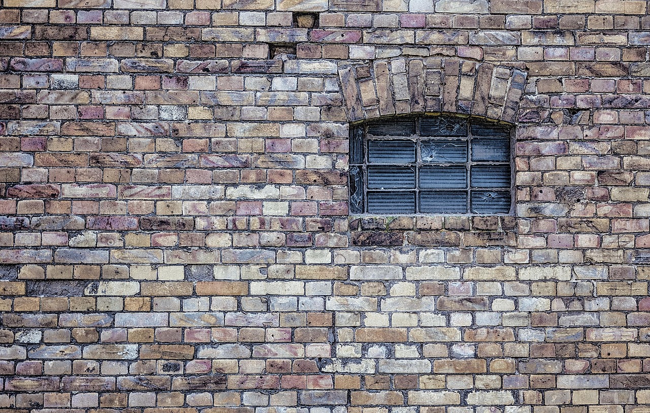 A flat brick wall in mottled yellow, pink and grey, with a small arched window set into it, its metal grille divided into nine rectangular panes