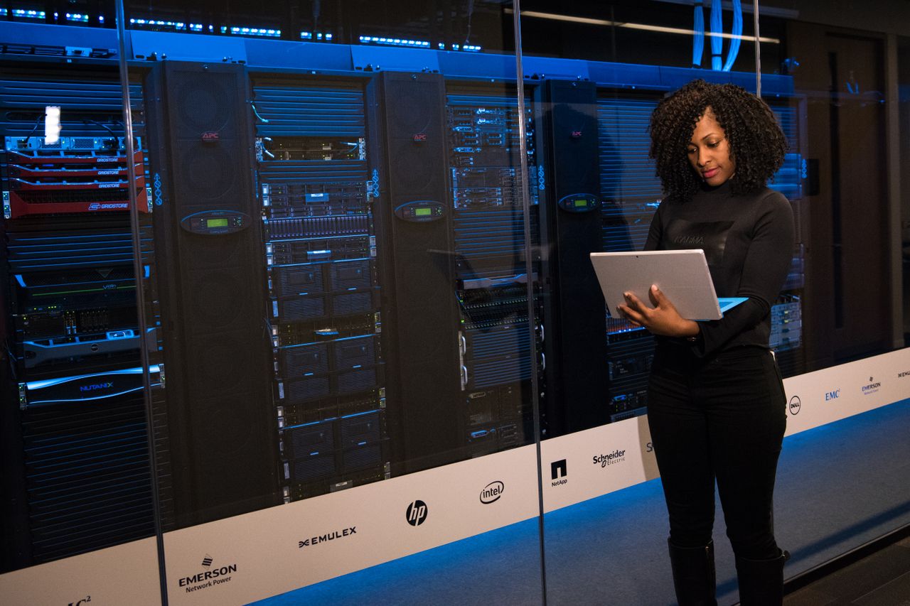 A person standing in front of glass-walled server racks in a data center, holding an open laptop, with blue lighting and vendor logos visible on the glass panels