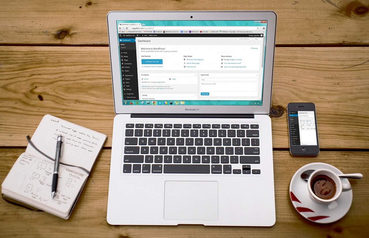 A MacBook Air on a wooden desk showing the WordPress admin dashboard, beside a phone, notebook and coffee