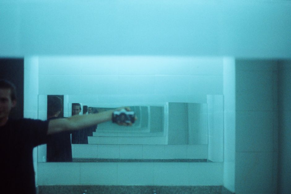 A person photographing themselves in a bathroom mirror facing another mirror, so the reflection repeats into the distance in ever smaller frames, the whole scene tinted blue-green