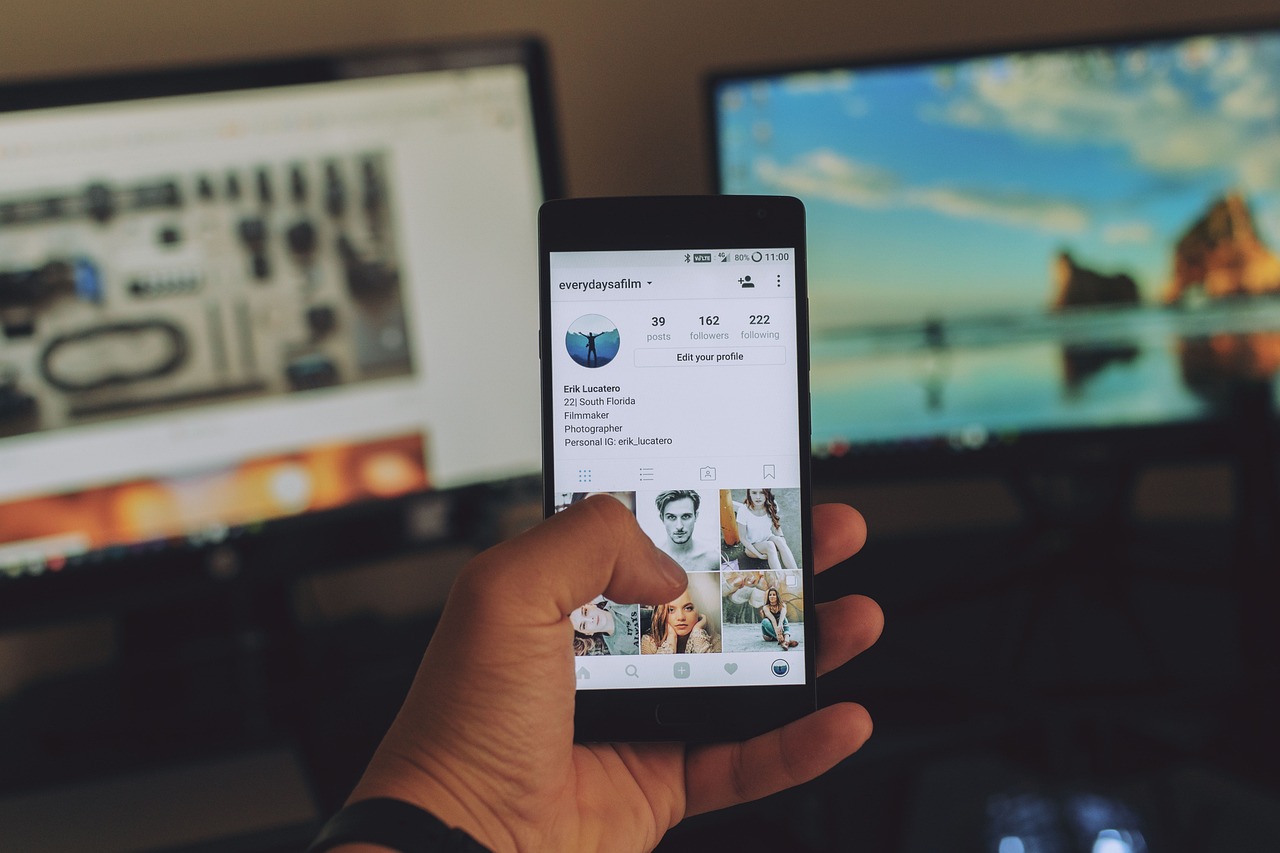 A hand holding a phone showing an app profile screen in front of two desktop monitors