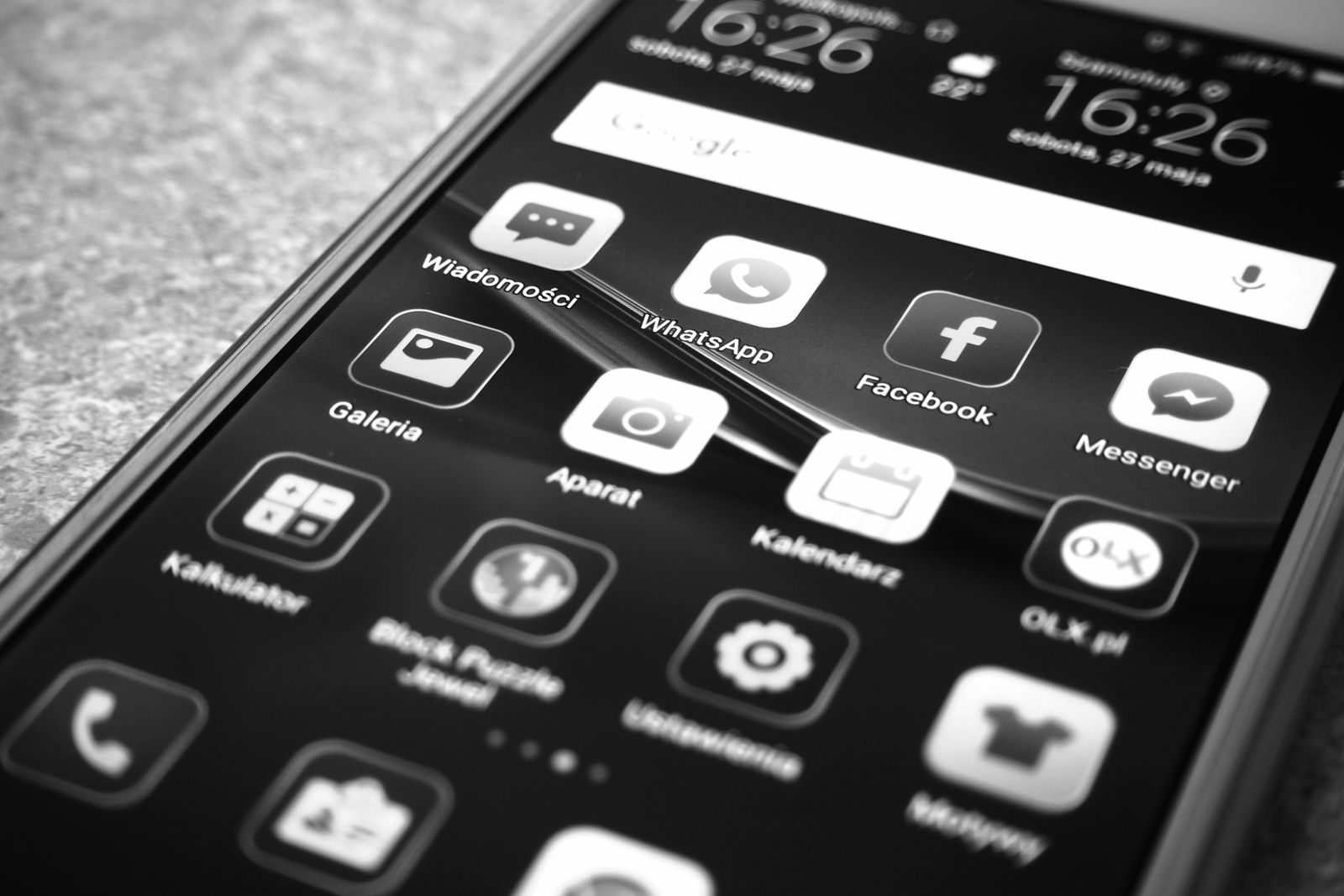 Black and white close-up of an Android phone lying at an angle on a speckled surface, its home screen showing a Google search bar, a clock reading 16:26 on Saturday 27 May, and app icons labelled in Polish including Wiadomosci, WhatsApp, Facebook, Messenger, Galeria, Aparat and Kalendarz