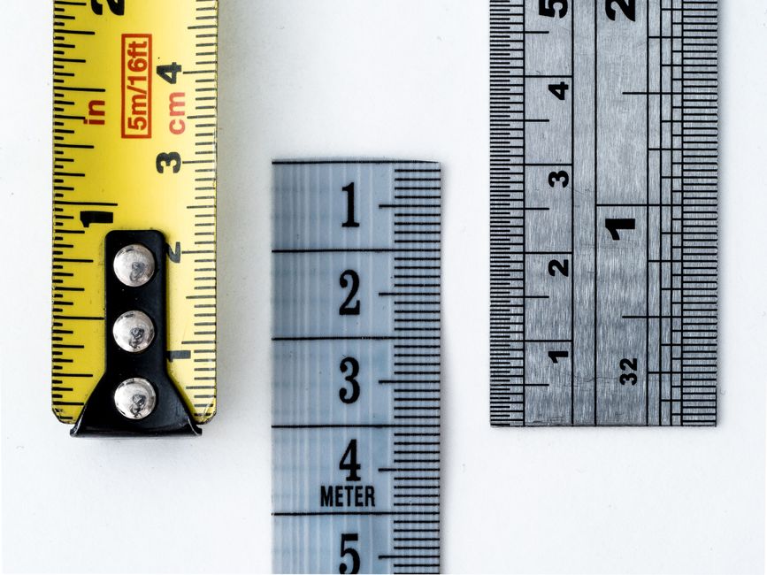 Three measuring instruments photographed from above on a white surface: a yellow tape measure marked 5m/16ft in inches and centimetres, a translucent plastic ruler marked METER, and a steel rule with fine graduations