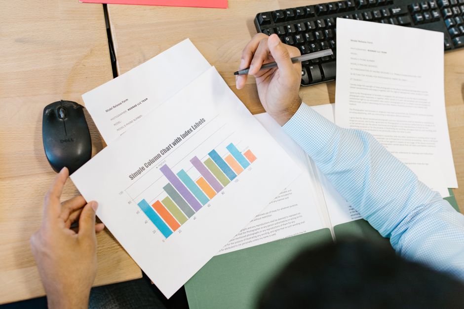 Overhead view of a desk: a hand holding a pen above a printed sheet showing a colour bar chart titled Simple Column Chart with Index Labels, next to a mouse, a keyboard and other printed forms