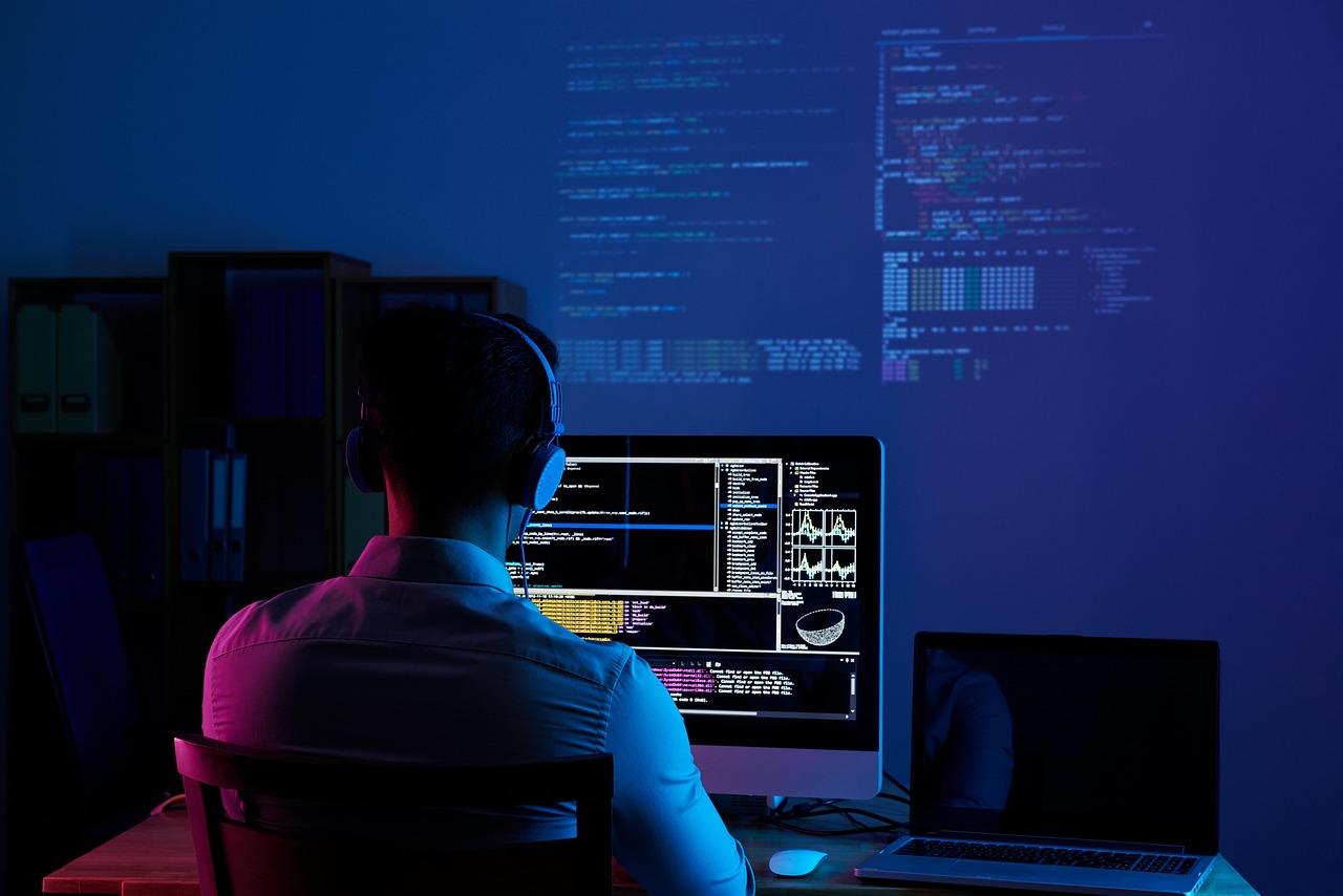 Un desarrollador con auriculares está sentado de espaldas a la cámara, frente a un monitor que muestra código en una sala con luz azul oscura
